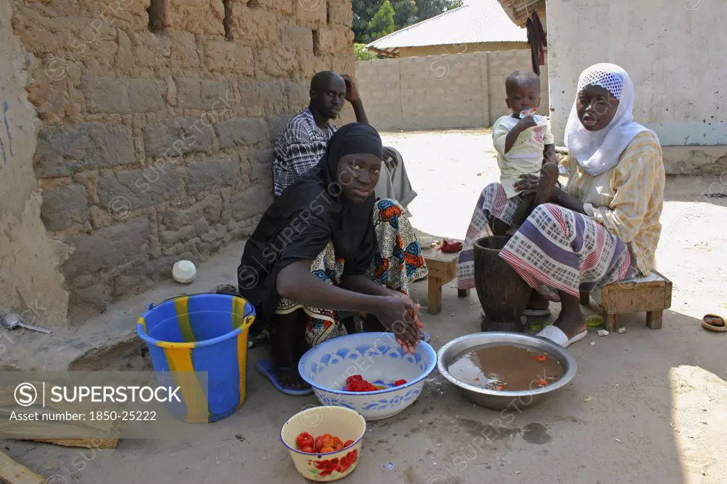Gambia, Western Gambia, Tanji, Man Sitting In Shade In Yard Outside His House In The Company Of Two Of His Wives Who Are Taking Care Of A Child While Washing And Smashing Tomatoes To Be Used In The Family'S Meal