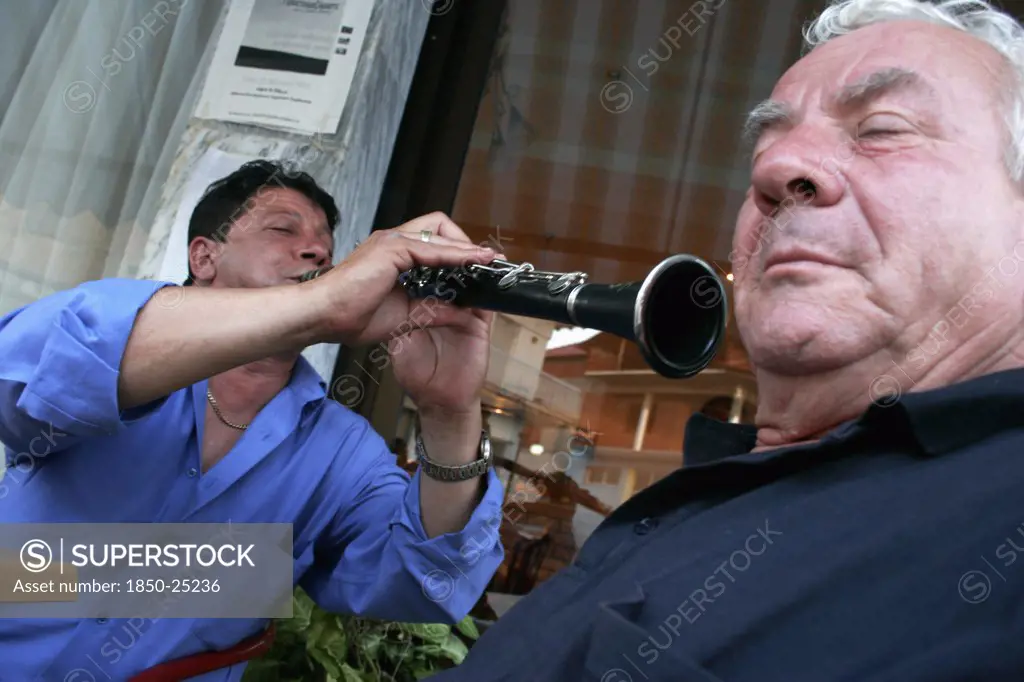 Greece, Macedonia, Kilkis, 'Toumba.  Street Musician Playing Clarinet, Perspective And Angle Of Picture Giving Appearance That He Is Directing Instrument And Sound Towards Ear Of Elderly Man In Foreground. '