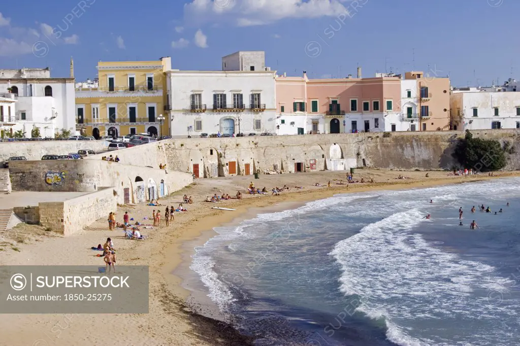 Italy, Puglia, Gallipoli, View Of Old City With Traditional Houses Beside Sea Wall Overlooking Curving Sandy Beach With People Sunbathing And Swimming