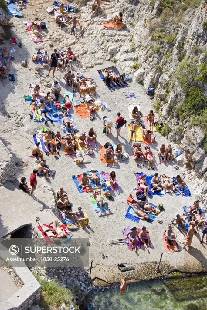 Italy, Puglia, Lecce, Tricase.  Looking Down On People Sunbathing On Coloured Beach Towels Laid Out On Rocks Next To The Sea.