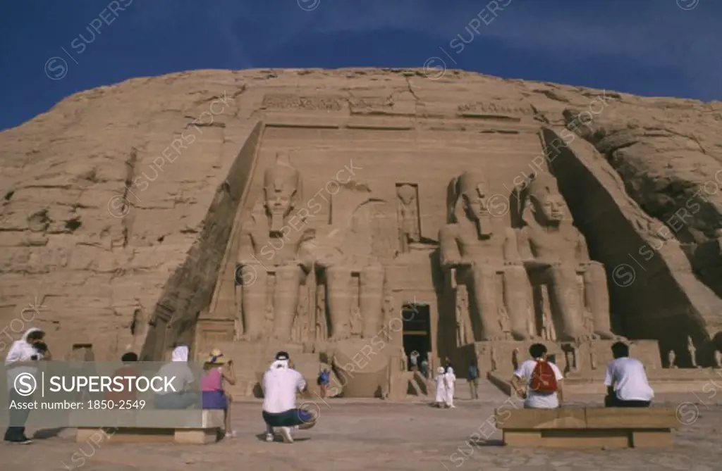 Egypt, Abu Simbel, Tourists Sitting In Front Of Giant Seated Statues Of Ramesses Ii At Temple Entrance