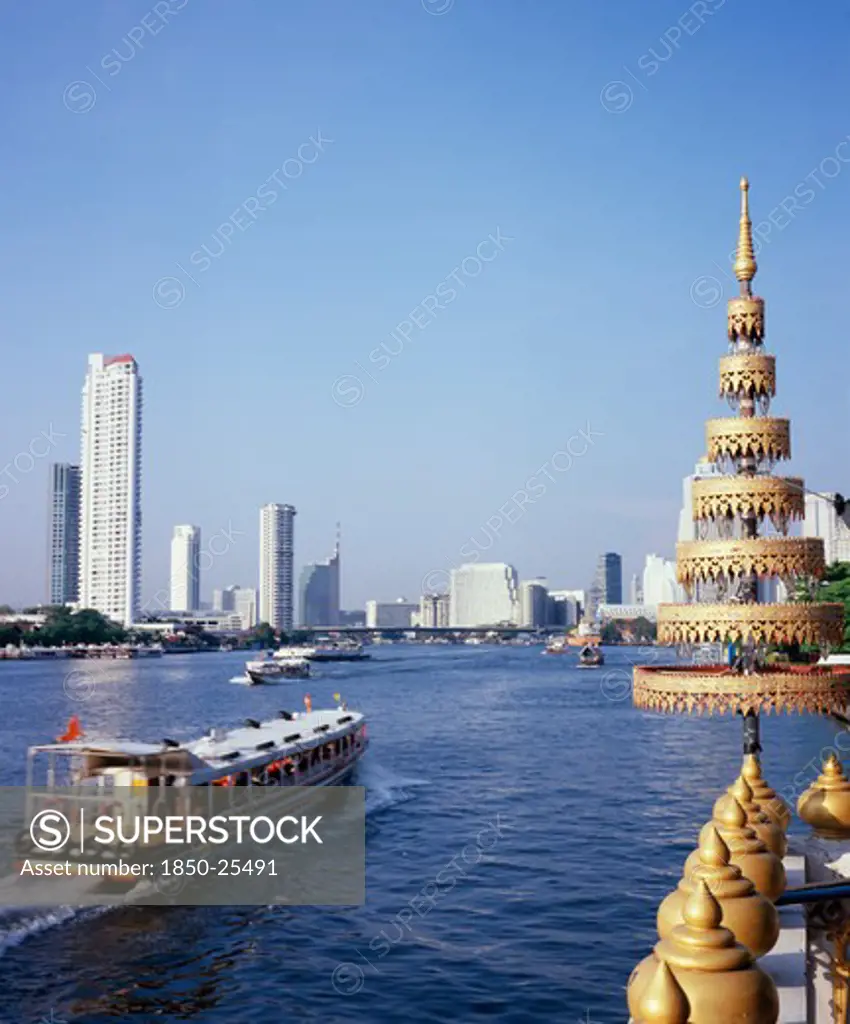 Thailand, Bangkok, 'View Across Chao Phraya River From Wat Ratchasungkhon Decorated With Gold Umbrella Usually Sheltering Buddha In Foreground With Express River Ferry, Indicated By Orange Flag Departing North For Saphan Thaksin Bridge Stop.  New Apartments, Shangri-La Hotel And High Rise Buildings On Opposite Bank.'