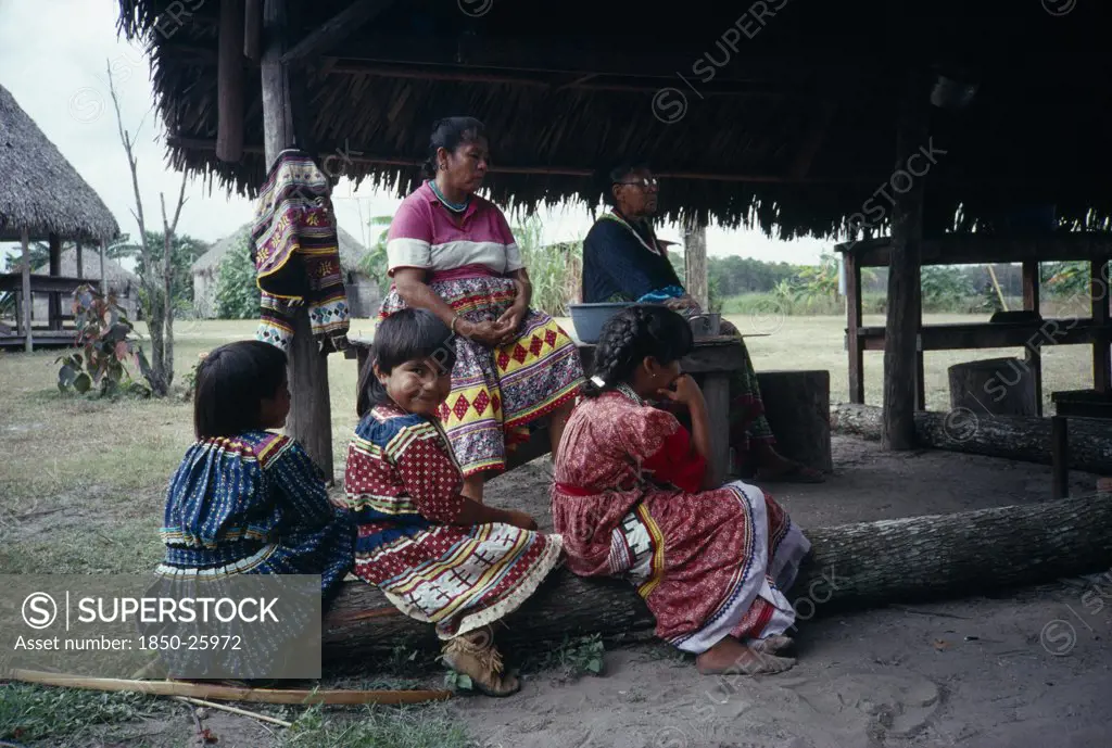 Usa, Florida, Everglades, Independent Seminole Native American Village. Family Of Women And Young Girls Wearing Traditional Colourful Patchwork Dresses Sitting Under A Chickee Hut.