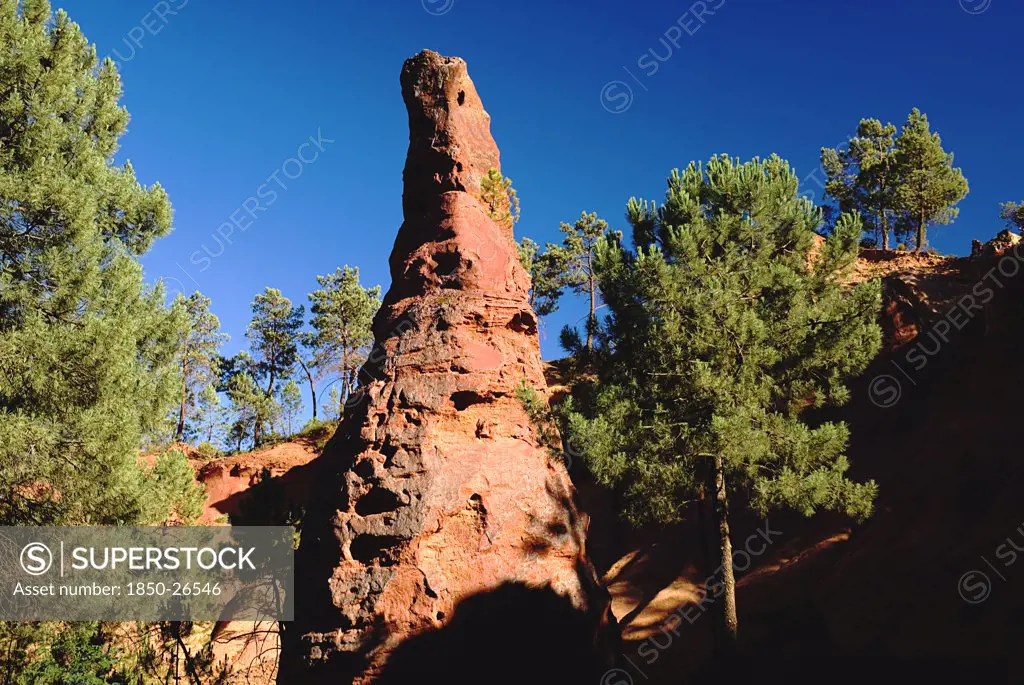 France, Provence Cote DAzur, Le Sentier Des Ocres (The Ochre Footpath) , 'A Tall Thin, Eroded Rock Pinnacle In The Area Known As The Needle Cirque Part Circled By Trees Against Cloudless Blue Sky.'