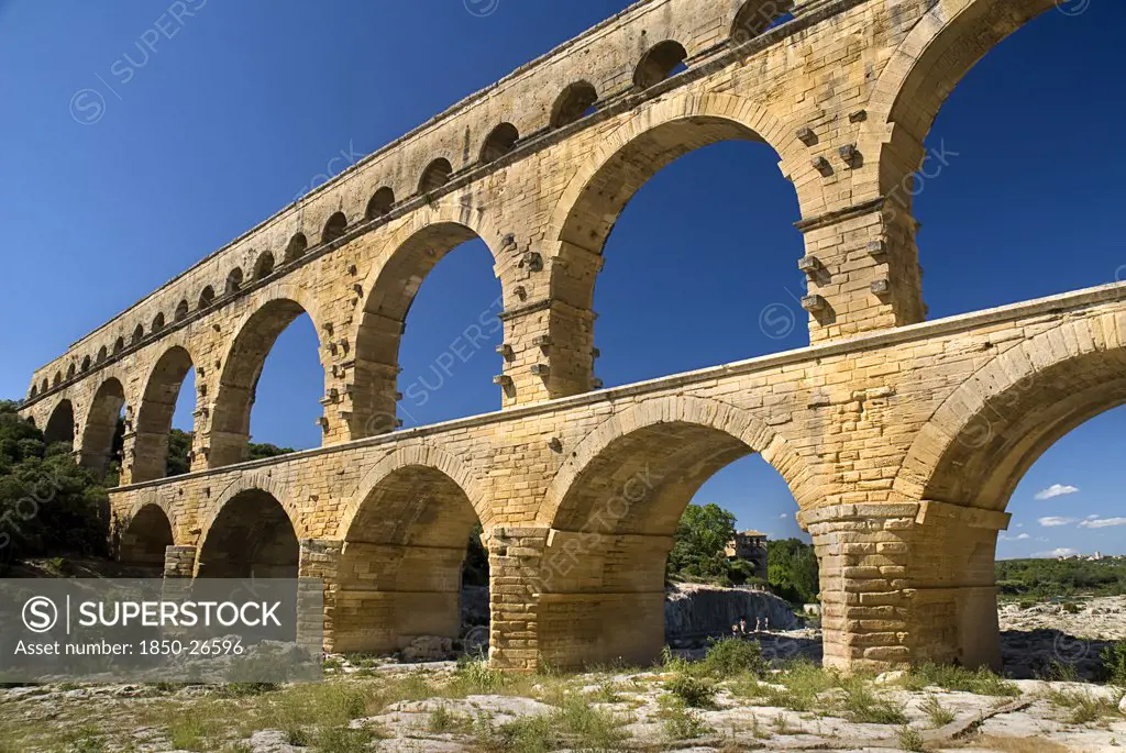 France, Provence Cote DAzur, Gard, Pont Du Gard.  Angled View Of Roman Aqueduct From The West Side In Glowing Evening Light Showing Three Tiers Of Continuous Arches Against Cloudless Blue Sky.