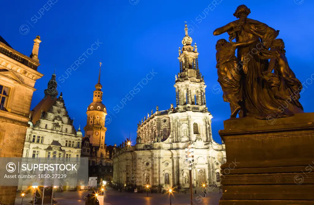 Germany, Saxony, Dresden, The 18Th Century Hofkirche Catholic Cathedral Of Saint Trinitatis And The Illuminated Tower Of The Residenzschloss And The Grunes Gewelbe Or Green Vault A Museum That Is Part Of Dresden Castle With A Statue Of A Woman And Children In The Foreground At Sunset.