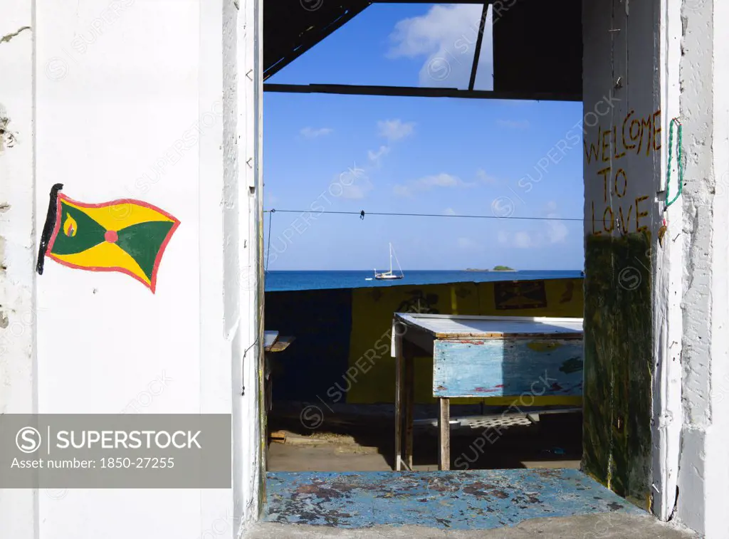 West Indies, Grenada, Carriacou, Hillsborough Derelict Building In The Main Street With The Grenadian Flag And Welcome To Love Painted On The Walls With A Yacht At Anchor In The Harbour Beyond The Ruined Building.