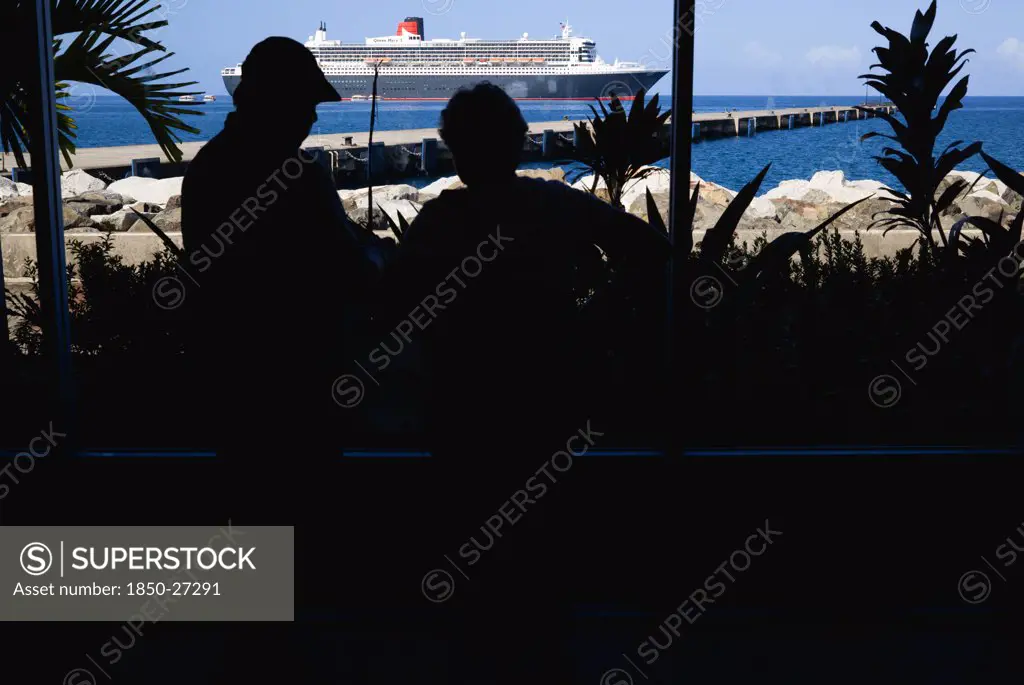 West Indies, Grenada, St Georges, Tourists In The Cruise Ship Terminal Looking Out Through The Glass Window Towards The Queen Mary 2 Liner Moored Offshore.
