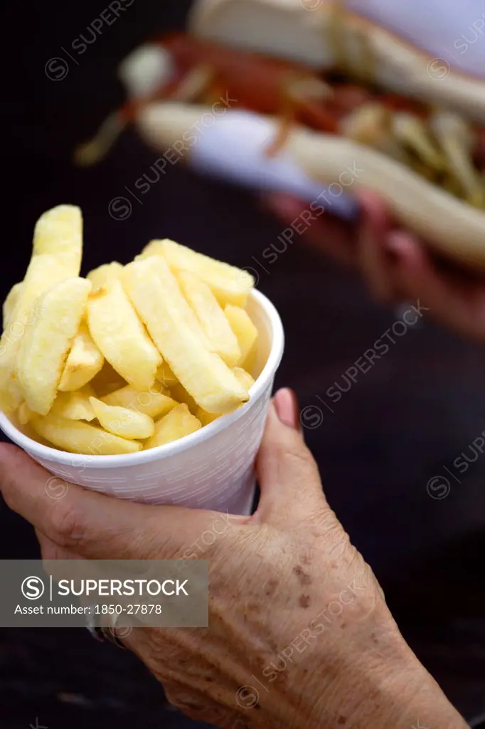 England, West Sussex, Findon, Findon Village Sheep Fair Elderly Lady Holding A Polystyrene Mug Full Of Potato Fries In One Hand And A Hot Dog With Tomato Ketchup And Onions In The Other.