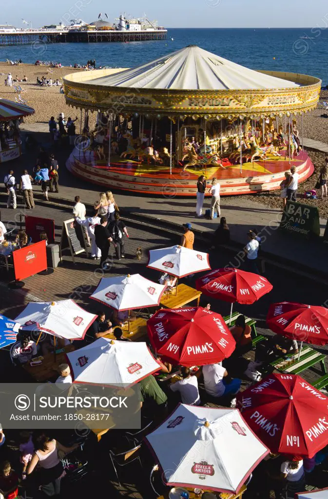 England, East Sussex, Brighton, People Sitting Under Sun Shade Umbrellas At Tables On The Promenade Outside Bars And Restaurants With A Traditional Victorian Galloping Horses Carousel Fairground Roundabout And Brighton Pier With People On The Shingle Pebble Beach Beyond.