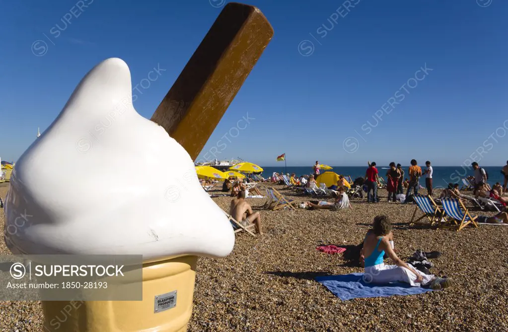 England, East Sussex, Brighton, People Sunbathing On The Pebble Shingle Beach With A Giant Fibreglass Model Of An Ice Cream Cone In The Foreground.