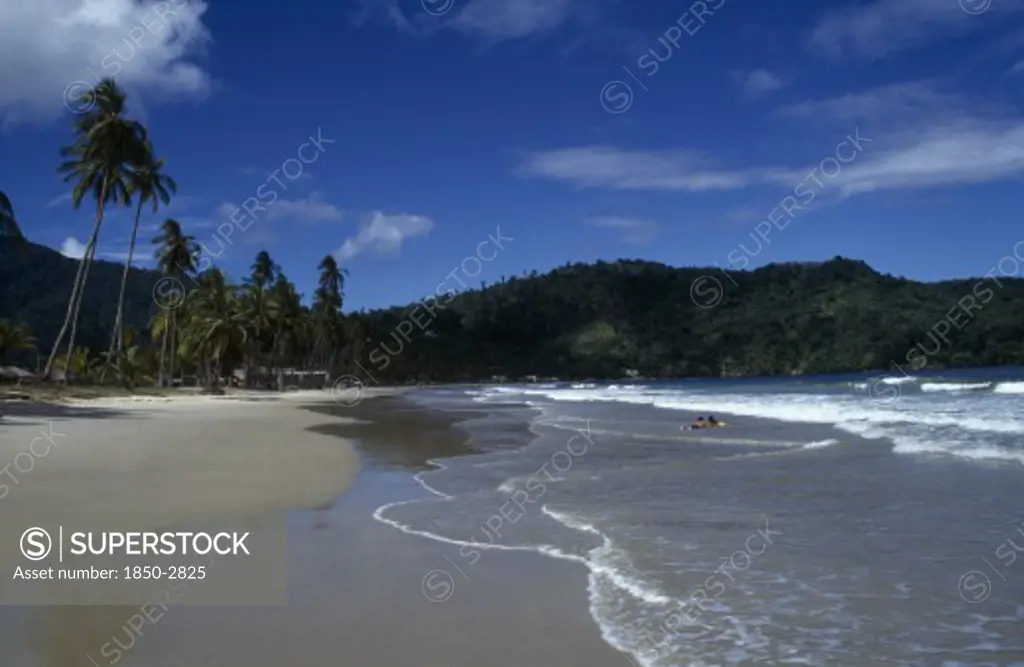 West Indies  , Trinidad, Maracas Bay , Sandy Beach With Couple Lying In The Surf And Tree Covered Headland Beyond.