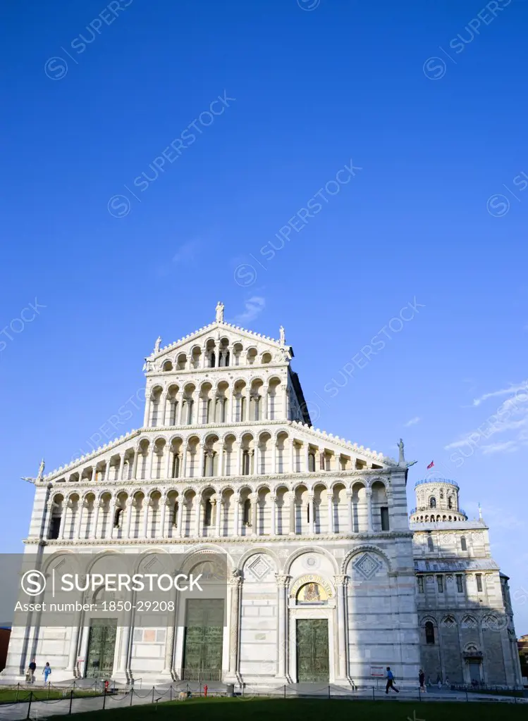 Italy, Tuscany, Pisa, The Campo Dei Miracoli Or Field Of Miracles With The Lombard Style 12Th Century Facade Of The Duomo Cathedral Church And The Leaning Tower Beyond Under A Blue Sky.