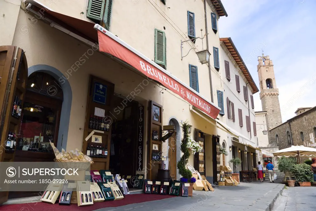 Italy, Tuscany, Montalcino, Val Dorcia Brunello Di Montalcino Enoteca Or Wine Shop With Display Of Boxed Wines On The Pavement Around Its Entrance Beneath A Sunshade. The 14 The Century Belltower Of The Palazzo Comunale Is At The End Of The Street.
