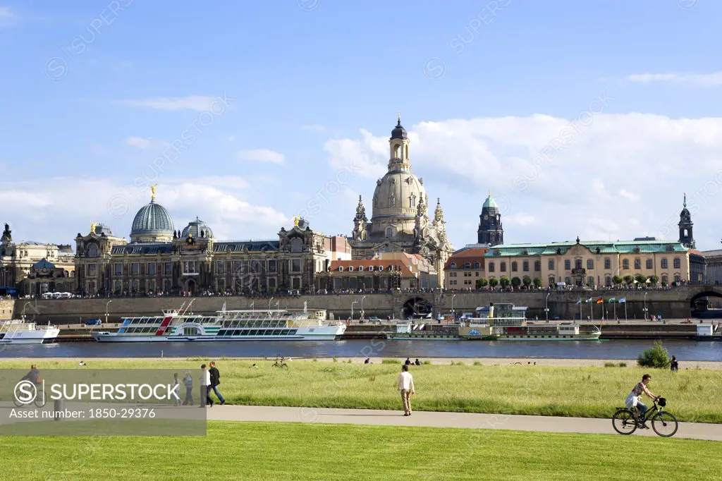 Germany, Saxony, Dresden, The City Skyline With Cruise Boats Moored On The River Elbe In Front Of The Embankment Buildings On The Bruhl Terrace Busy With Tourists Of The Art Academy The Restored Baroque Frauenkirche Church Of Our Lady Dome With Some People Sitting On The Grass Or Cycling Or Walking On The Path Of The Near River Embankment.