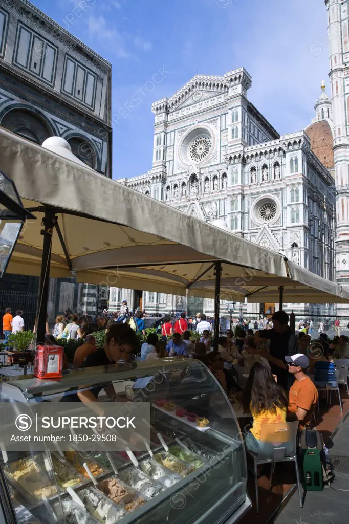 Italy, Tuscany, Florence, The Neo-Gothic Marble West Facade Of The Cathedral Of Santa Maria Del Fiore The Duomo With Sightseeing Tourists People Sitting Under Umbrellas In The Shade At A Restaurant And A Waitress Serving Ice Cream From A Counter.