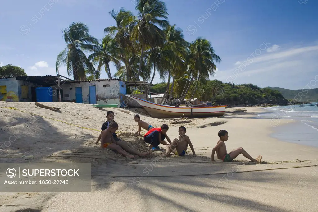 Venezuela, Margarita Island, Playa La Galera, Kids Playing On The Beach  Under The Shade Of A Palm Tree Just In Front Of The Seawater While Other Trees  A House And A Boat Are Noticeable At The Background  Shoot On A Bright Day With Blue Sky And Some White Clouds.