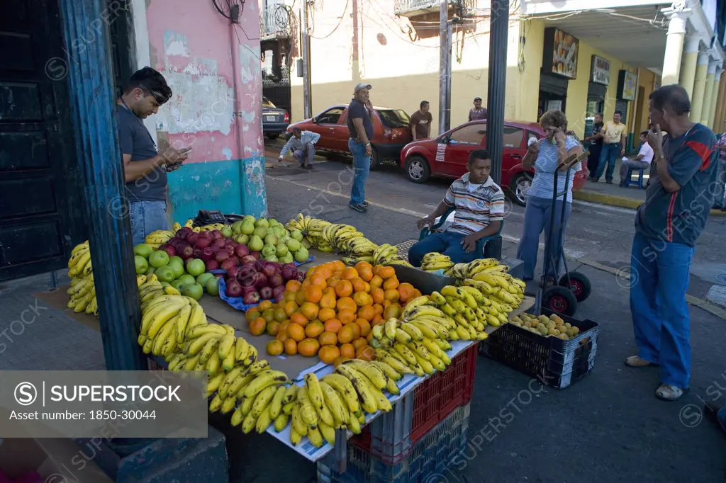 Venezuela, Bolivar State, Ciudad Bolivar, Fruit Stall With Bananas  Oranges  Apples And Other Fruits At Ciudad Bolivars Main Street Just By The Orinoco River With People Looking At Them And Talking To The Salesman While Other People  Cars And Old Buildings With Columns Are Noticeable At The Background. People Using Mobile Phones.