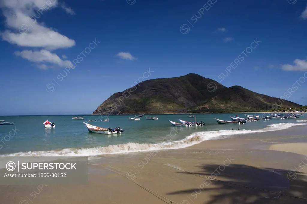 Venezuela, Margarita Island, Playa La Galera, View Of Exotic Beach With Palm Trees And Their Shades On The Sand  Just In Front Of Fishing Boats Floating At The Tropical Crystal Clear Seawaters  Shoot On A Bright Day With Blue Sky And White Clouds.