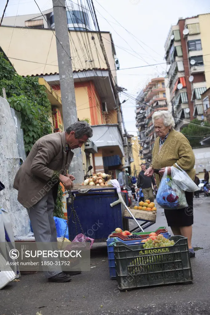 Albania, Tirane, Tirana, A street vendor using hand held set of scales to weigh fruit for elderly woman customer carrying bags.