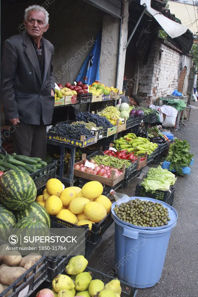 Albania, Tirane, Tirana, Grocer beside his shop front display of fruit and vegetables including melons  potatoes and grapes in the Avni Rustemi market.