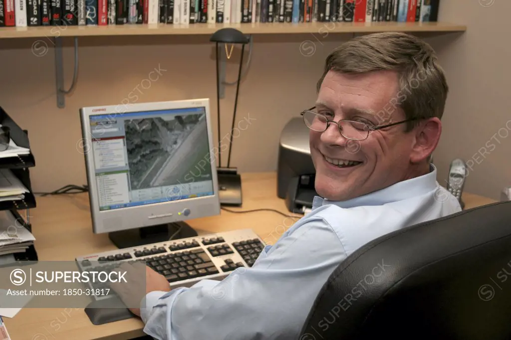 Media, Computer, Office, A man working from his home office  turning towards camera  sitting at desk and using PC. Printer  phone  filing tray of paperwork and shelf of books visible.