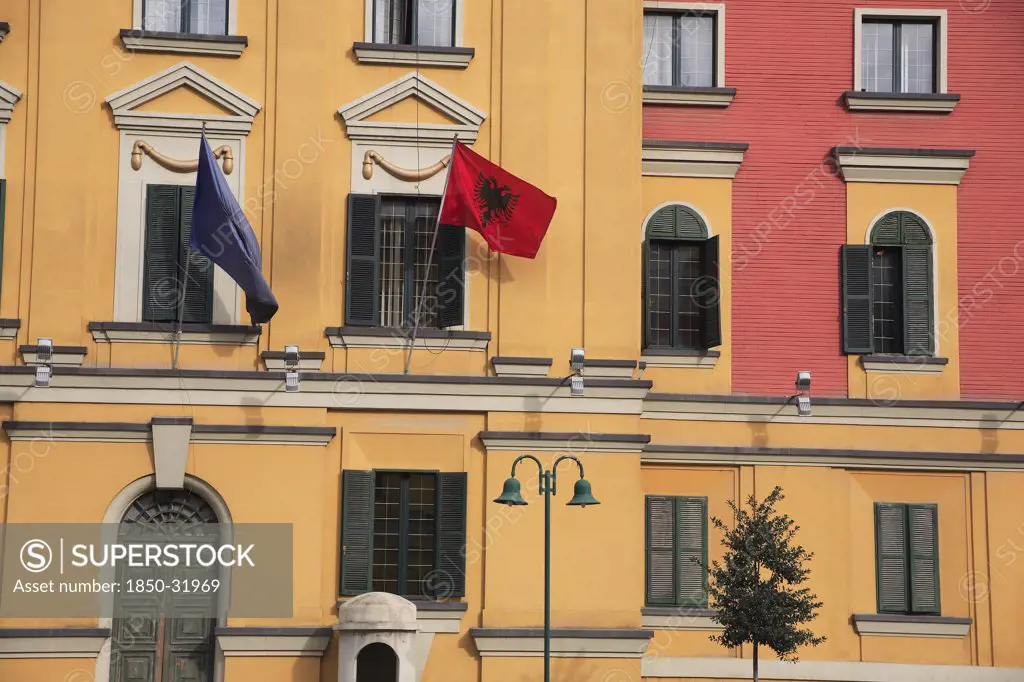 Albania, Tirane, Tirana, Part view of pink and yellow exterior facade of government buildings in Skanderbeg Square flying flags including double headed eagle emblem. Multiple windows with green painted shutters and green painted  arched doorway.
