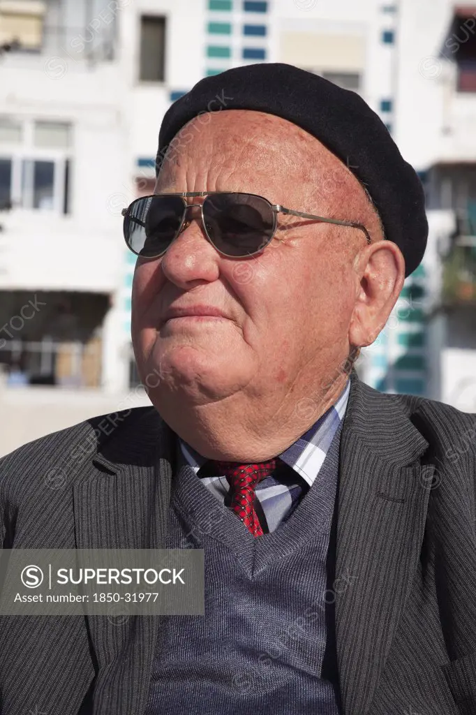 Albania, Tirane, Tirana, Head and shoulders portrait of an elderly man wearing sunglasses and a beret. Three-quarter profile left.