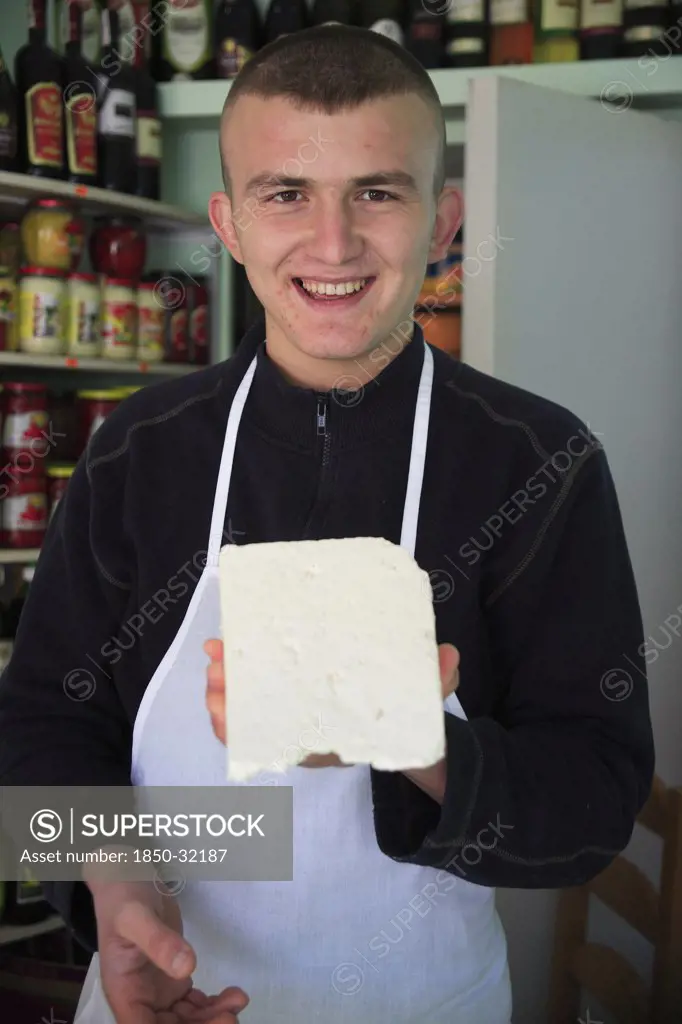 Albania, Tirane, Tirana, Young male cheese shop vendor in the Avni Rustemi Market wearing white apron and holding up block of cheese.