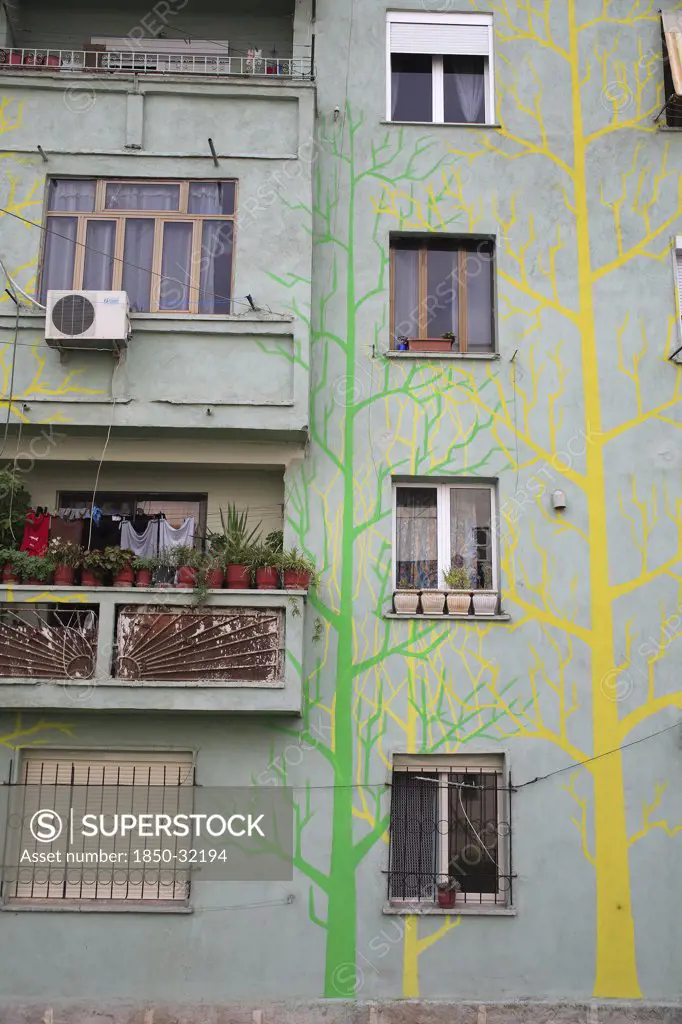 Albania, Tirane, Tirana, Part view of exterior facade of apartment block painted with tree forms in green and yellow  with washing hanging on balcony crowded with pot plants.