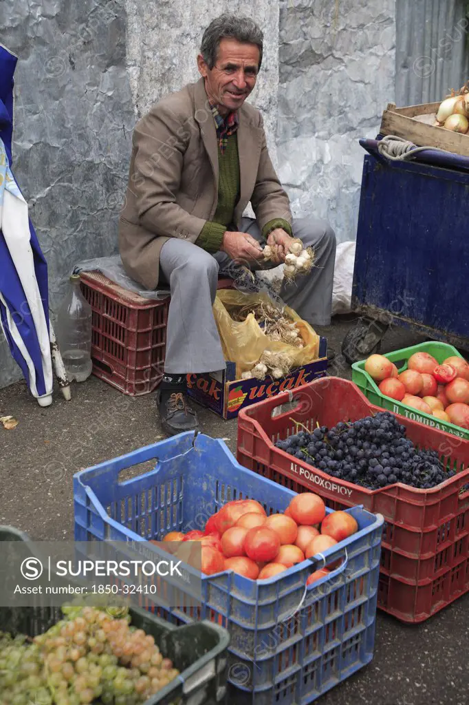 Albania, Tirane, Tirana, Fruit and vegetable street vendor in the Avni Rustemi Market seated on crate behind display including grapes and tomatoes.