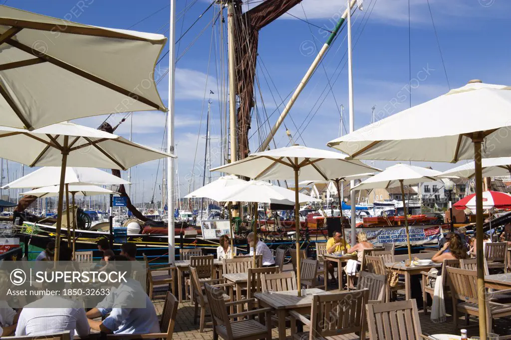 England, Hampshire, Portsmouth, Port Solent People at restaurant tables under sun shade umbrellas with yachts moored in the marina and housing beyond.