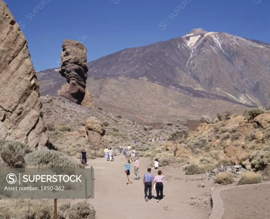 Spain, Canary Islands, Tenerife, Mount Teide National Park With Tourists On Dry And Dusty Path Amongst Rock Formations With Teide In The Distance