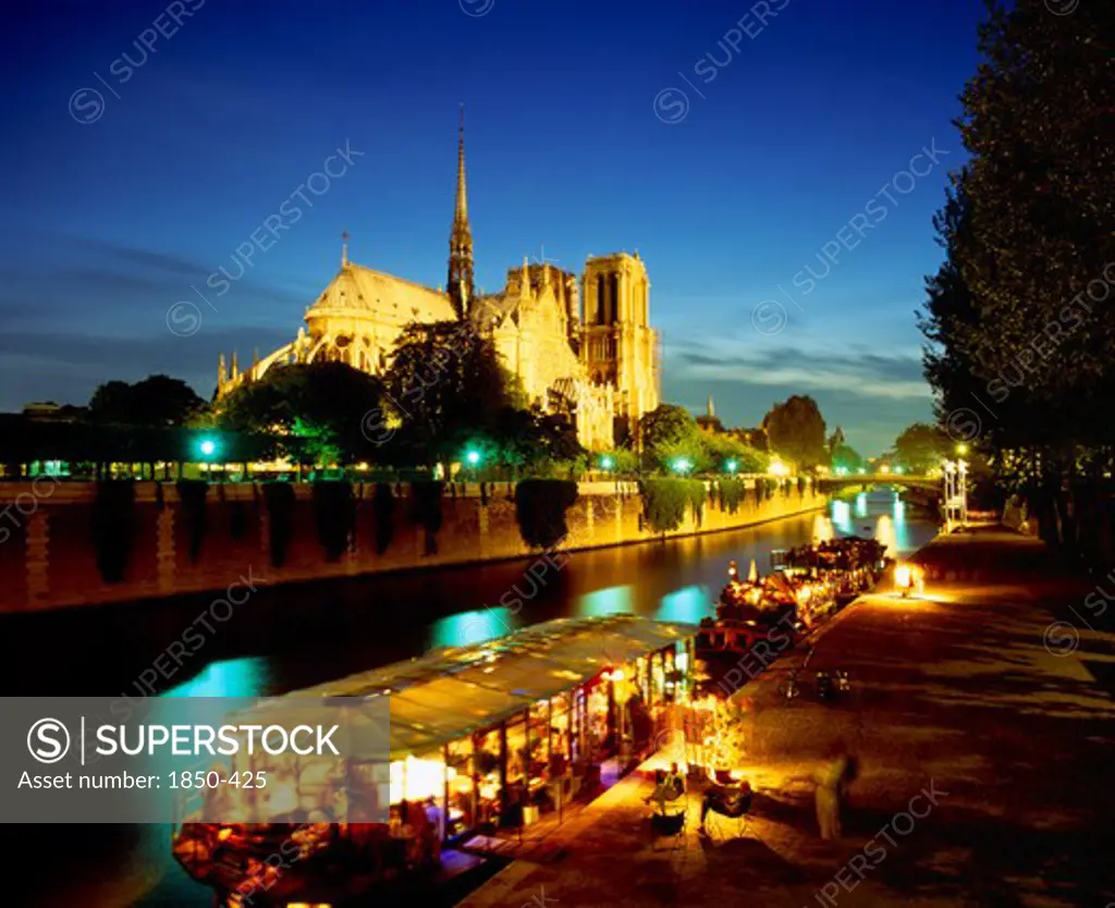 France, Ile De France, Paris, Notre Dame Floodlit At Night With Restaurant And Houseboat Lit Up On The River Seine In Foreground.