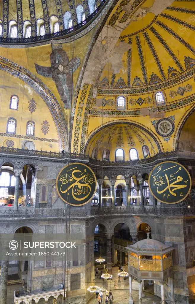 Turkey,  Istanbul, Sultanahmet Haghia Sophia Sighseeing tourists beneath the dome with murals and chandeliers in the Nave of the Cathedral with calligraphic roundels of Arabic Koran texts abobe the Lodge of The Sultan.