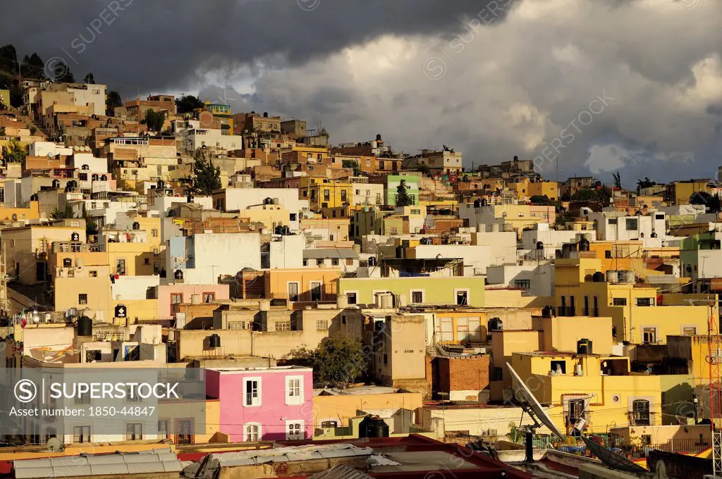 Colourful houses clinging to the hillside below sky of thick grey cloud. , Mexico Bajio Zacatecas