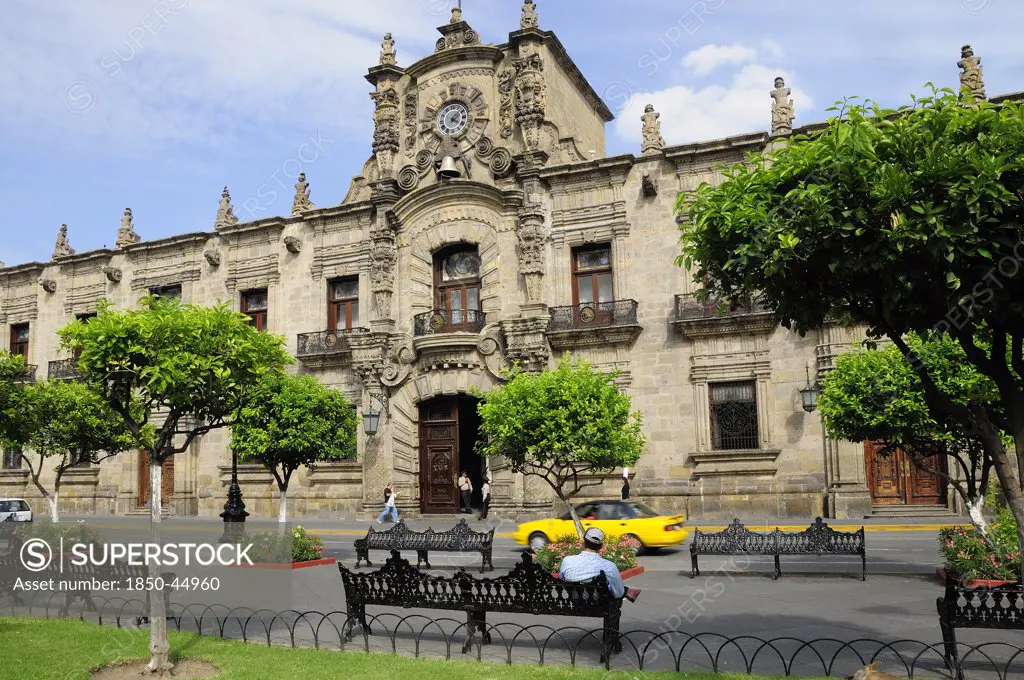 Palacio Gobierno the Government Palace. Exterior facade from Plaza de Armas with passing car trees and man seated on bench in foreground, Mexico Jalisco Guadalajara