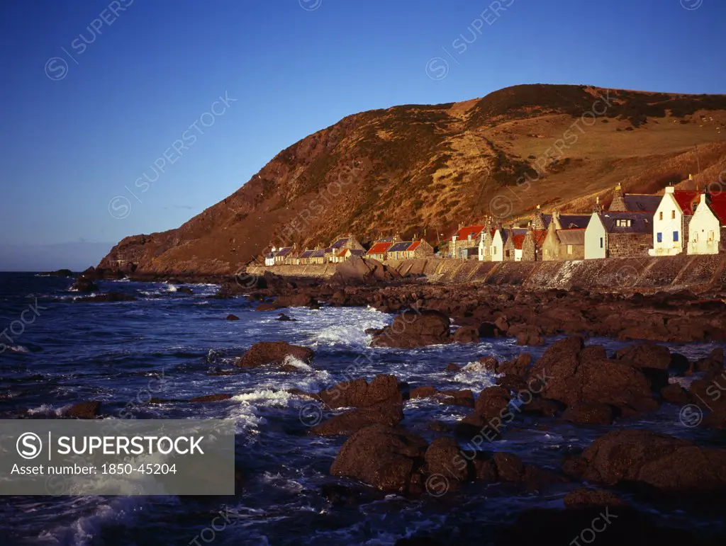 One time fishing village seen from shoreline. Row of cottages at foot of steep hillside overlooking coast and stone jetty.Scotland Aberdeenshire Crovie