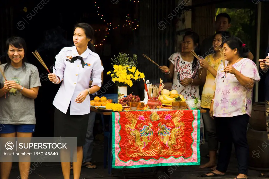 Thailand, Bangkok, Woman in military uniform holding joss sticks beside offerings on decorated table celebrating local temple parade.