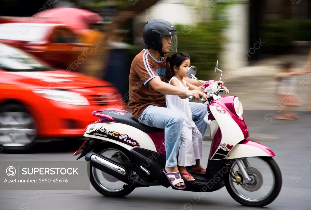 Thailand, Bangkok, Father with young daughter on motorcycle pass brightly coloured taxis.
