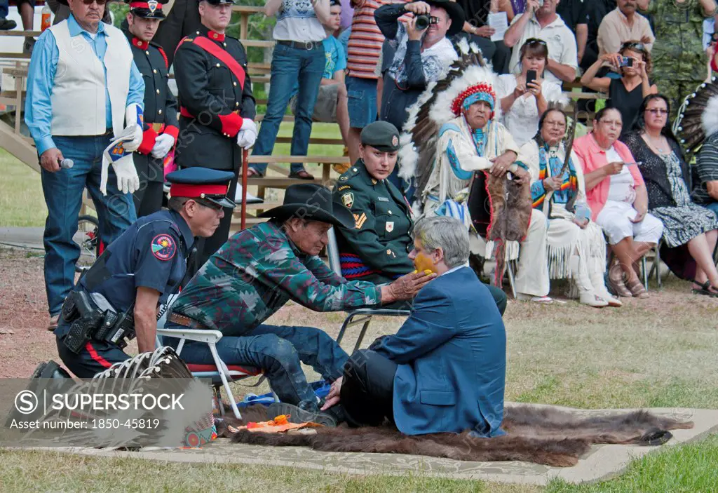 Canada, Alberta, Stand Off, Canada's Prime Minister, Stephen Harper, seated on a buffalo skin rug as Medicine Man Pete Standing Alone applies paint to his face as part of the ceremony to induct him into Kainai or Blood Tribe Chieftainship, Blood Tribe Police, Blood Tribe Master Corporal in the Canadian Forces wounded in Afghanistan, Chiefs andi Elders look on.