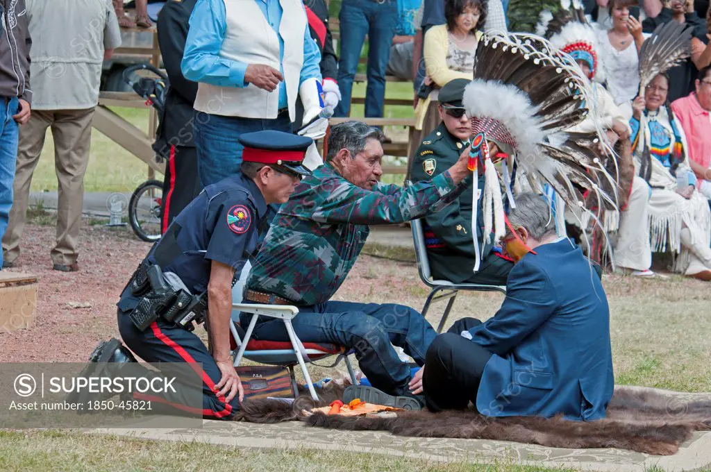 Canada, Alberta, Stand Off, Canada's Prime Minister, Stephen Harper, seated on a buffalo skin rug as Medicine Man Pete Standing Alone places a ceremonial  headdress of Eagle feathers on his head as part of the ceremony to induct him into Kainai or Blood Tribe Chieftanship, He was given the Kainai or Blood Tribe name Chief Speaker, Blood Tribe Police, Chiefs and Elders look on.