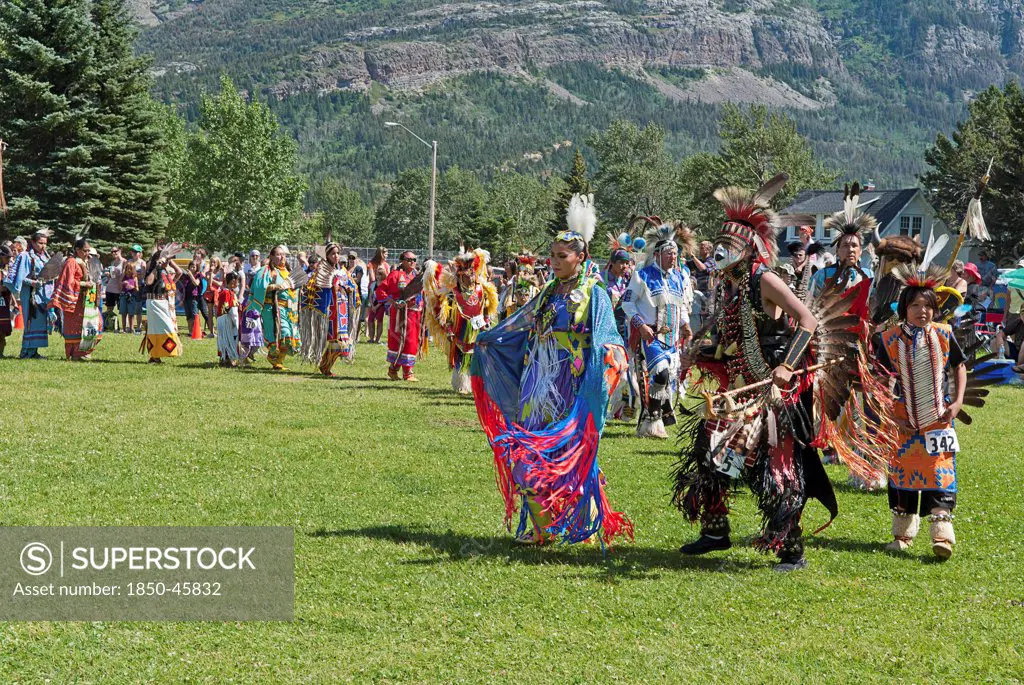 Canada, Alberta, Waterton Lakes National Park, Grand Entry of dancers in full regalia at the Blackfoot Arts & Heritage Festival Pow Wow organized by Parks Canada and the Blackfoot Canadian Cultural Society at this UNESCO World Heritage Site, Mount Vimy in the background, Green grass and trees.