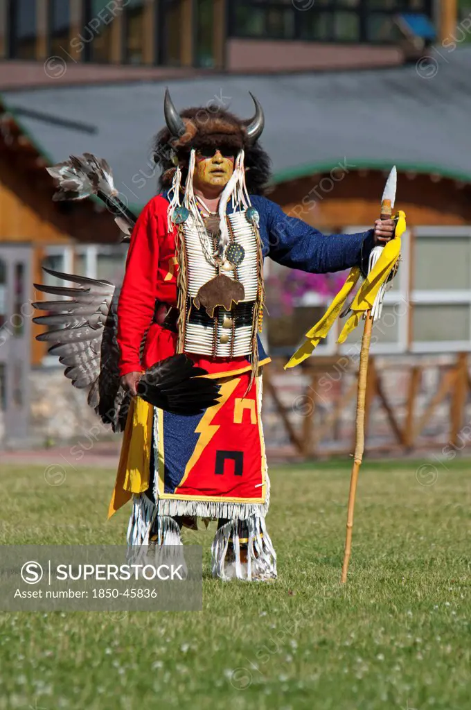 Canada, Alberta, Waterton Lakes National Park, Blackfoot dancer in a buffalo headdress and sunglasses wearing a porcupine quill breastplate and holding a spear and feather fan at the Blackfoot Arts & Heritage Festival Pow Wow organized by Parks Canada and the Blackfoot Canadian Cultural Society at this UNESCO World Heritage Site red, blue yellow, green, complementary colours.