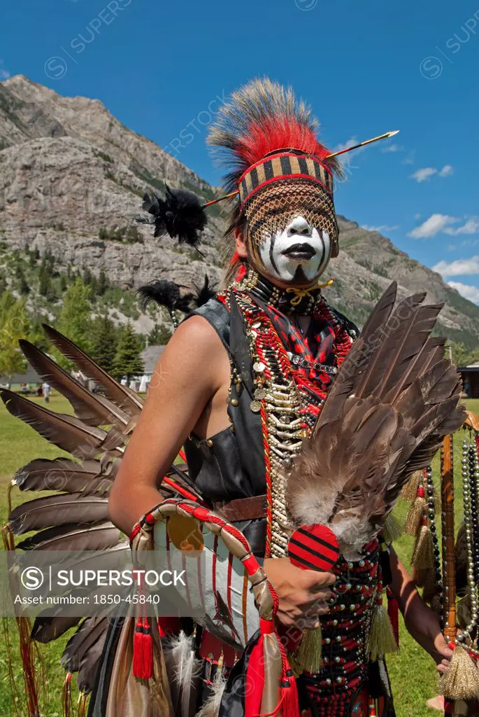Canada, Alberta, Waterton Lakes National Park, Blackfoot head dancer Aryson Black Plume in full regalia and face paint at the Blackfoot Arts & Heritage Festival Pow Wow organized by Parks Canada and the Blackfoot Canadian Cultural Society, Headdress with arrow and black plume, Feather fan and bustle, Rocky Mountains blue sky with white clouds, red, blue, green, dominant red, UNESCO World Heritage Site.