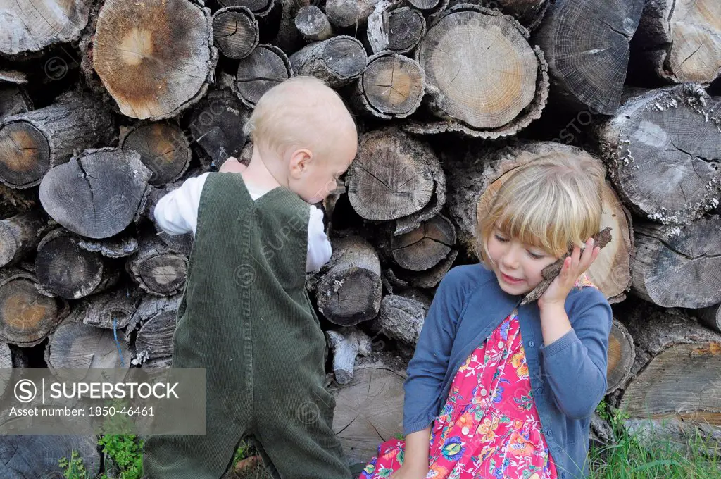 Kids, Outdoor, 5 year old Eva and Oscar aged two, playing beside a log pile with Eva using bark as pretend mobile phone.