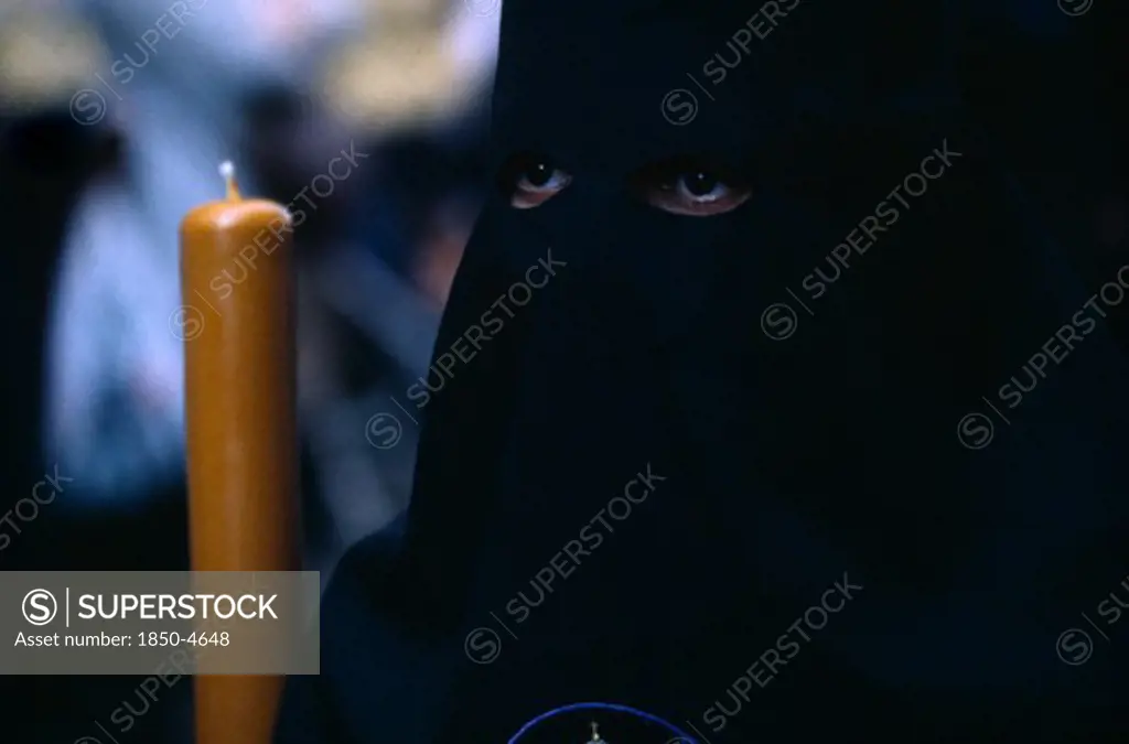 Spain, Andalucia, Seville, Semana Santa Easter Procession. Penitent In Black Robe With Eyes Visable Through Hood Holding A Candle