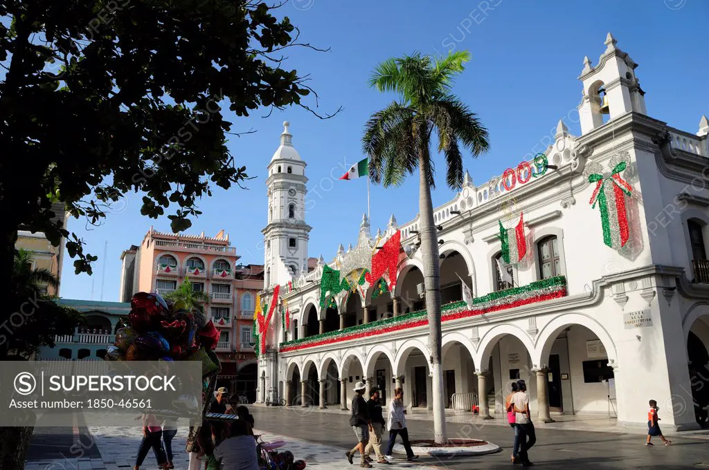 Mexico, Veracruz, The zocalo and government buildings decorated for Independence Day celebrations with balloon seller and tourists in the foreground part framed by tree.