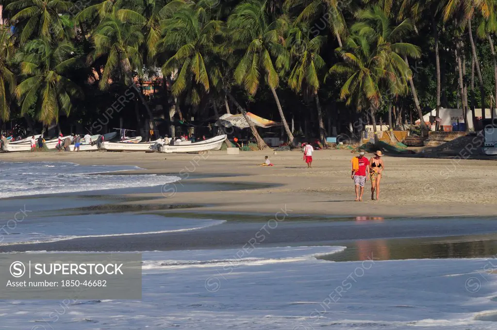 Mexico, Oaxaca, Puerto Escondido, Playa Marinera with couple walking along shore line of boats pulled up onto sand and overhanging palms beyond.