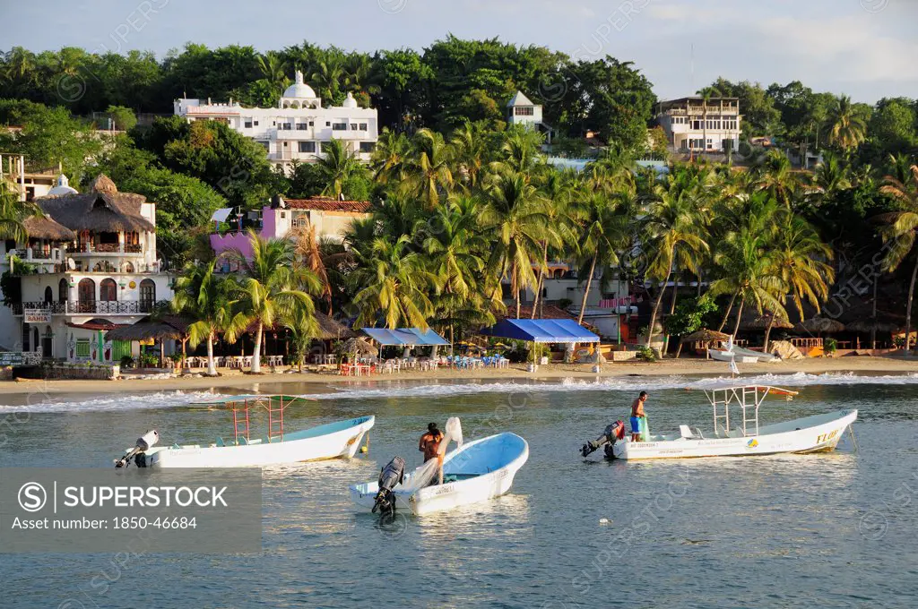 Mexico, Oaxaca, Puerto Escondido, Fishing boats in bay at Playa Marinero lined with cafes bars and other buildings amongst palm trees.